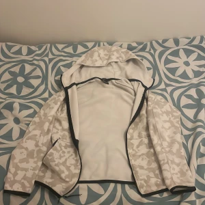 Nike camo vindjacka med huva - Snygg vit och beige camo vindjacka från Nike med svart dragkedja och huva. Jackan har två fickor framtill och är perfekt för dig som gillar streetstyle. Tillverkad i lätt polyester och har lång ärm. Nike-logga på bröstet för extra stilpoäng.