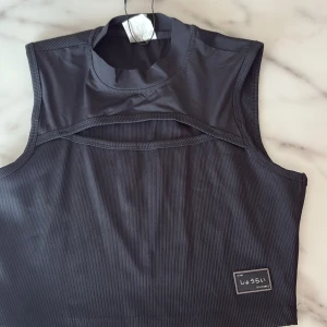 Svart croppad ribbad linnetopp - Svart croppad linnetopp med hög hals och cool cut-out detalj framtill. Toppen är ribbad och har en liten patch med japansk text nere till höger. Perfekt för dig som gillar en edgy och modern stil.