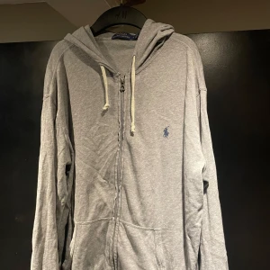 Grå hoodie från Polo Ralph Lauren - Snygg grå hoodie från Polo Ralph Lauren med dragkedja och huva. Tröjan har vita snören, känguruficka och den klassiska blå loggan broderad på bröstet. Tillverkad i mjuk bomull, perfekt för chill dagar eller när du vill ha en clean look.