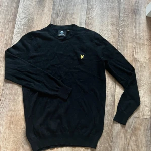 Svart stickad tröja från Lyle & Scott - Svart stickad tröja från Lyle & Scott med rund hals och klassisk gul logga på bröstet. Tröjan har lång ärm och ribbade muddar vid ärmslut och nederkant. Perfekt för dig som gillar stilren och enkel design.