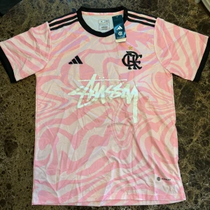Adidas x Stüssy Flamengo rosa tröja M - Unik fotbollströja från Adidas i samarbete med Stüssy och Flamengo. Rosa och vit med swirl-mönster, svart krage och ärmslut. Stüssy-logga tryckt på bröstet, Flamengo-emblem och Adidas-märke. Tillverkad i lätt polyester med Aeroready-teknologi.
