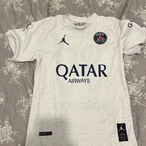 PSG Messi 30 vit matchtröja Jordan - Snygg PSG fotbollströja i vitt med mörkblå detaljer, Messi 30 på ryggen och Qatar Airways-tryck på bröstet. Tröjan har korta ärmar, rund hals och är tillverkad i lätt och ventilerande material. PSG-emblem och Jordan-logga framtill. Perfekt för fans!