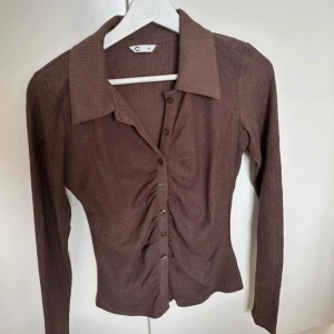 Brun skjorta med knappar och rynk - Snygg brun skjorta med långa ärmar och markerad krage. Skjortan har knappar framtill och cool rynkdetalj längs mitten som ger en trendig look. Perfekt att styla med jeans eller kjol för en chill vibe.