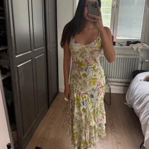 Blommig långklänning med volanger - Superfin långklänning med smala axelband och v-ringning. Klänningen är vit med blommigt mönster i grönt, rosa, blått och gult. Den har flera volanglager nedtill som ger en flowy känsla. Perfekt för sommarens alla tillfällen!