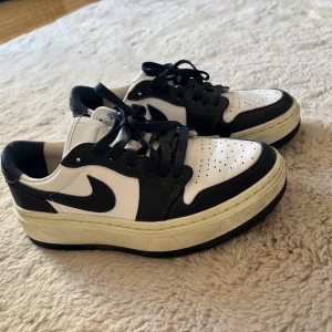 Nike Jordan 1 Low svart/vit - Snygga Nike Jordan 1 Low sneakers i svart och vitt skinn med klassisk swoosh på sidan. Skorna har svarta snören, perforerad tå och en platt sula. Perfekta för dig som gillar streetstyle och vill ha en ikonisk look.