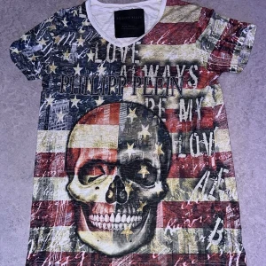 Philipp Plein t-shirt med USA-flagga - Snygg t-shirt från Philipp Plein med tryck inspirerat av amerikanska flaggan, stjärnor och ränder i rött, vitt och blått. Stora logotypen framtill och coola textdetaljer. Rund halsringning med vit kant. Perfekt för dig som gillar statement-plagg.