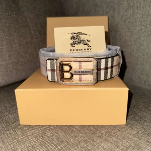 Burberry rutig bälte med B-spänne - Snyggt bälte från Burberry med klassiskt rutigt mönster i beige, svart, rött och vitt. Bältet har ett stort guldfärgat B-spänne med Burberry-logga. Perfekt accessoar för att lyfta din outfit med en lyxig touch.