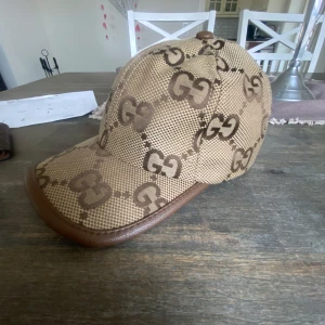 Beige och brun keps från Gucci - Snygg keps från Gucci i beige och brunt med det ikoniska GG-mönstret. Kepsen har en justerbar rem i brunt skinn baktill för perfekt passform. Perfekt för att ge din outfit en lyxig touch.
