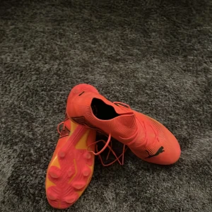 Röda fotbollsskor från Puma - Snygga röda fotbollsskor från Puma med svart logga. Skorna har snörning och en strömlinjeformad design för optimal prestanda på planen. Perfekta för dig som vill sticka ut med stil. Storlek 43. Skorna är nya endast använda en gång.