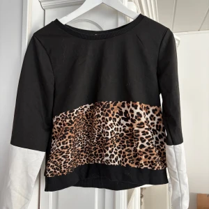 Svart tröja med leopardmönster - Snygg svart tröja med leopardmönstrad detalj i mitten och vita ärmar. Tröjan har en ribbad kant vid halsen. Perfekt för en trendig look!