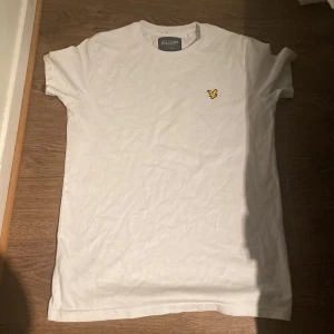 Vit t-shirt från Lyle & Scott - Säljer en klassisk vit t-shirt från Lyle & Scott med deras ikoniska gula logotyp på bröstet. T-shirten är kortärmad och tillverkad i mjukt bomullsmaterial. Perfekt för en stilren och avslappnad look.