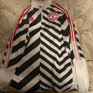Svartvit randig fotbollströja från Adidas Manchester United  - Säljer en unik svartvit randig fotbollströja från Adidas med Manchester United-logga på bröstet. Tröjan har röda detaljer och en halv dragkedja framtill. Perfekt för fans av laget!