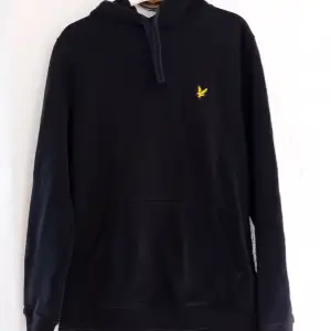 Säljer en svart hoodie med gul Lyle&Scott logga på bröstet. Tröjan har en klassisk design med huva och en stor ficka framtill. Perfekt för en avslappnad stil.