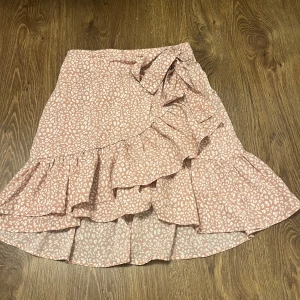 Rosa mönstrad kjol från Shein - Säljer en söt rosa kjol från Shein med ett ljust mönster och volangdetaljer. Kjolen har en omlott-design och är perfekt för en somrig look. Den är gjord i ett lätt och luftigt material.