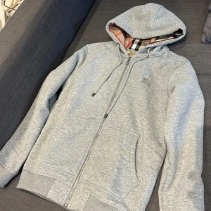 Grå huvtröja från Burberry - Säljer en stilren grå huvtröja från Burberry med dragkedja och klassiskt rutigt mönster i huvan. Tröjan har långa ärmar och ribbade muddar vid ärmslut och nederkant. Perfekt för en avslappnad look.