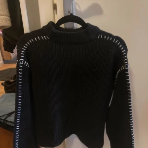 Svart stickad tröja med vita detaljer - Säljer en svart stickad tröja med vita sömmar längs ärmarna och axlarna. Använd men inga slitningar. Inget nopprigt material