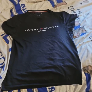 Svart t-shirt från Tommy Hilfiger - Säljer en svart t-shirt från Tommy Hilfiger med tryck på bröstet. Tillverkad i ekologisk bomull för en mjuk och bekväm känsla. Perfekt för en stilren och avslappnad look.