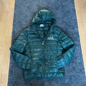 EA7 pufferjacka  - Säljer en snygg grön pufferjacka från EA7. Jackan har en dragkedja framtill och en praktisk huva. Perfekt för kyligare dagar och ger en sportig look. Passar dig mellan 160-172