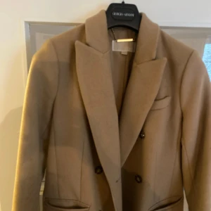 Beige kappa från Michael Kors - Elegant beige kappa från Michael Kors med dubbelknäppning och klassisk krage. Kappan har långa ärmar med dekorativa knappar vid ärmsluten och fickor framtill. Perfekt för en stilren look.