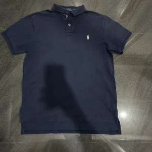 Mörkblå pikétröja från Ralph Lauren - Säljer en klassisk mörkblå pikétröja från Ralph Lauren med den ikoniska loggan broderad på bröstet. Tröjan har en krage och knappar framtill. Perfekt för en stilren och avslappnad look.