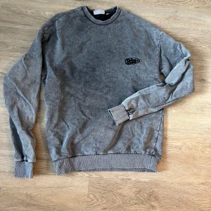 Grå sweatshirt från Junkyard - Säljer en grå sweatshirt från Junkyard med ett litet svart broderat märke på bröstet. Tröjan har långa ärmar och ribbade muddar vid ärmslut och nederkant. Perfekt för en avslappnad stil.