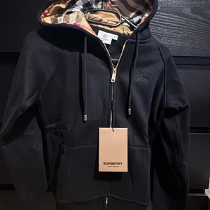 Burberry kofta  - Säljer en stilren svart huvtröja från Burberry med dragkedja och klassiskt rutigt foder i huvan. Tröjan har långa ärmar och en praktisk ficka framtill. Perfekt för en avslappnad och trendig look.