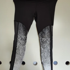 Svarta leggings med vit mönsterdetalj - Snygga svarta leggings från H&M med en cool vit mönsterdetalj som sträcker sig från knäna och neråt. Perfekta för träning eller en avslappnad dag. Tillverkade i ett stretchigt syntetmaterial för bästa komfort.