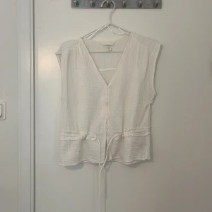 Vit ärmlös blus med knyt - Säljer en stilren vit blus med v-ringning och knytband i midjan. Blusen är ärmlös och har en luftig design, perfekt för varma dagar. 