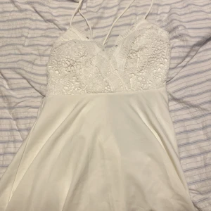 Vit klänning med spetsdetaljer - Säljer en söt vit klänning med spetsdetaljer på överdelen och tunna axelband. Klänningen har en v-ringad design och är perfekt för sommarens varma dagar. Den är kort och har en luftig passform.