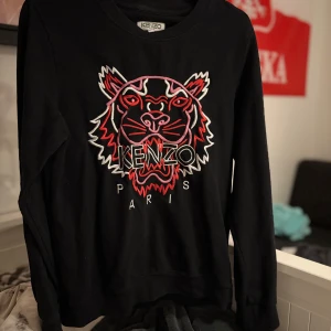 Kenzo tröja med tigerbroderi - Säljer en svart tröja från Kenzo med ett coolt tigerbroderi i rött, vitt och rosa på framsidan. Säljer den pga att den legat i en flyttlåda och skräpat i 3-4 år. Ordinarie pris ligger på ca 2200 kr. Kan eventuellt gå ner lite i pris vid snabb affär och seriös köpare.