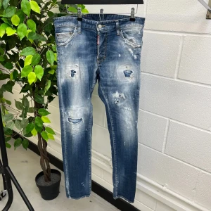 Blå jeans från DSQUARED2 - Säljer ett par coola blå jeans från DSQUARED2 med slitna detaljer och en vintage look. Jeansen har en klassisk femficksdesign och en röd logolapp bak. Perfekta för en avslappnad stil.