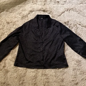 Svart leopardmönstrad blus från Bik Bok - Snygg svart blus från Bik Bok med ett diskret leopardmönster. Blusen har en klassisk krage och knäppning framtill. Perfekt för att ge din outfit en trendig touch.