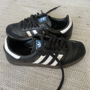 Svarta Adidas Samba sneakers - Säljer ett par klassiska svarta Adidas Samba sneakers med vita ränder och bruna sulor. Skorna har snörning och är perfekta för en sportig look. Använt ett par fåtal gånger! 