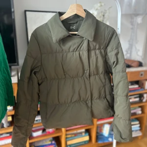 Olivgrön dunjacka från Uniqlo - Säljer en dunjacka från Uniqlo. Jackan har en stilren design med knappar framtill och en klassisk krage. Perfekt för kyligare dagar med sin quiltade stil och långa ärmar.