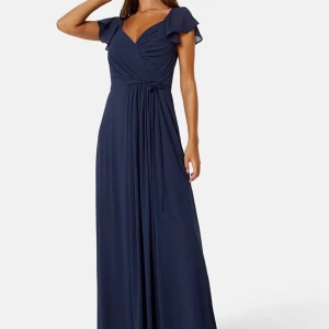 Marinblå långklänning med volangdetaljer - Elegant marinblå långklänning med omlott-design och volangdetaljer vid axlarna. Klänningen har en v-ringad hals och ett knytband i midjan för en smickrande passform. Perfekt för festliga tillfällen. Nypris 699kr