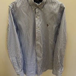 Randig skjorta från Polo Ralph Lauren - Säljer en klassisk blå- och vitrandig skjorta från Polo Ralph Lauren. Den är broderad med den ikoniska logotypen på bröstet. Perfekt för en stilren look.
