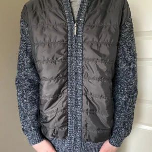 Grå fodrad cardigan med stickade ärmar - Snygg grå fodrad cardigan med quiltad framsida och stickade ärmar. Cardiganen har en dragkedja och en kamouflage mönstrad insida. Perfekt för lager-på-lager under kyliga dagar. Han på bilden är 180cm och väger 61kg. Inget märke tyvärr. Priset är diskuterbart