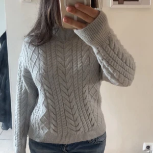 Grå stickad tröja från New yorker  - Säljer en stilren grå stickad tröja från New Yorker. Tröjan har ett snyggt flätmönster och en hög krage som ger extra värme. Perfekt för kyliga dagar. Den har långa ärmar och ribbade kanter för en bekväm passform. Passar utmärkt till både jeans och kjol.