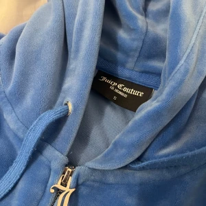 Blå velour hoodie från Juicy Couture - Säljer en mjuk och bekväm blå velour hoodie från Juicy Couture. Den har dragkedja framtill och en praktisk huva med snörning. Perfekt för en avslappnad stil. Priset kan diskuteras 