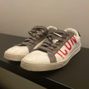 Vita sneakers från Dsquared2 med röd text - Snygga vita sneakers från Dsquared2 med stor röd text 'ICON' på sidan. Skorna har grå snörning och en svart detalj vid hälen. Perfekta för en stilren look.