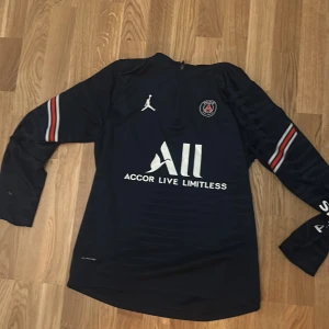 Mörkblå långärmad tröja från laget PSG  - Säljer en svart långärmad fotbollströja från Jordan med PSG-logga och texten 'Accor Live Limitless' på framsidan. Tröjan har vita och röda detaljer på ärmarna och texten 'Visit Rwanda' på baksidan. Perfekt för fotbollsfans!