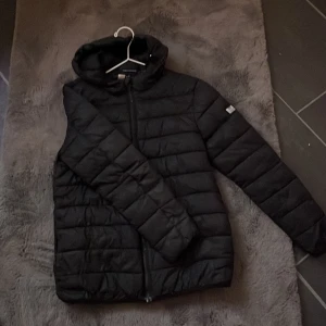 Svart pufferjacka från Weekend Offender - Säljer en stilren svart pufferjacka från Weekend Offender. Jackan har en dragkedja framtill och en praktisk huva. Perfekt för kyligare dagar och ger en modern look.