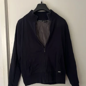 Svart kofta från Abacus - Säljer en stilren mörkblå kofta från Abacus med dragkedja och ribbad krage. Koftan har långa ärmar och en enkel design som passar perfekt för olika tillfällen. Perfekt för dig som gillar en klassisk look. Storleken är S-M.