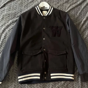 Unik Bomberjacka - Snygg svart bomberjacka från Wood Wood med vita ränder på krage, ärmslut och nederkant. Jackan har långa ärmar och knappar framtill samt ett broderat 'W' på bröstet. Perfekt för en stilren look.  Nypris ligger på  runt 2000kr enligt bildsökning.