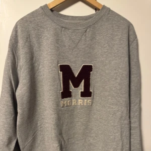 Grå sweatshirt från Morris - Säljer en stilren grå sweatshirt från Morris med ett stort broderat M på bröstet. Tröjan har långa ärmar och en klassisk rund halsringning. Perfekt för en avslappnad look.
