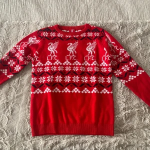 Röd jultröja med L.F.C. mönster - Säljer en röd jultröja från L.F.C. med vita och svarta detaljer. Tröjan har snöflingor, rutor och klubbens logga som mönster. Perfekt för unga Liverpool-fans som vill sticka ut under vintern och julen.