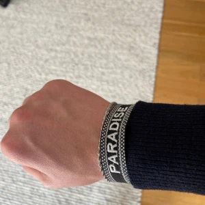 Svart och vitt armband med texten PARADISE - Säljer ett snyggt armband i svart och vitt med texten PARADISE. Armbandet har ett justerbart spänne. Köptes för 499kr men kan tänka 150kr
