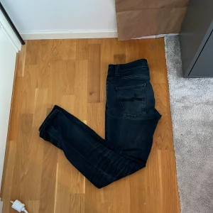 Mörkblå/svarta jeans från Nudie Jeans - Säljer ett par svarta jeans från Nudie Jeans, modell Grim Tim. Jeansen har klassisk femficksdesign, normal passform och raka ben. Perfekta för dig som gillar stilrena och tidlösa jeans. Kan väl vara värt att notera att det är två hål i vardera hörn i vänster ficka, men de är inte så stora och kan hålla mobilen.
