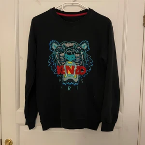 Svart sweatshirt med tigerbroderi från Kenzo - Svart sweatshirt från Kenzo med ikoniskt tigerbroderi i blått, grönt, rött och gult framtill samt texten 'KENZO PARIS'. Tröjan har rund halsringning och långa ärmar. Perfekt statement-plagg för dig som gillar streetwear.