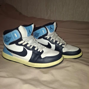 Nike Skor  i blått och vitt - Säljer ett par Nike skor med högt skaft i blått, vitt och svart. Skorna har klassisk snörning, svart swoosh och ljusblå detaljer runt ankeln och hälen. Perfekta för dig som gillar streetwear och vill sticka ut med snygga sneakers.! INTE RIKTIGA JORDANS!